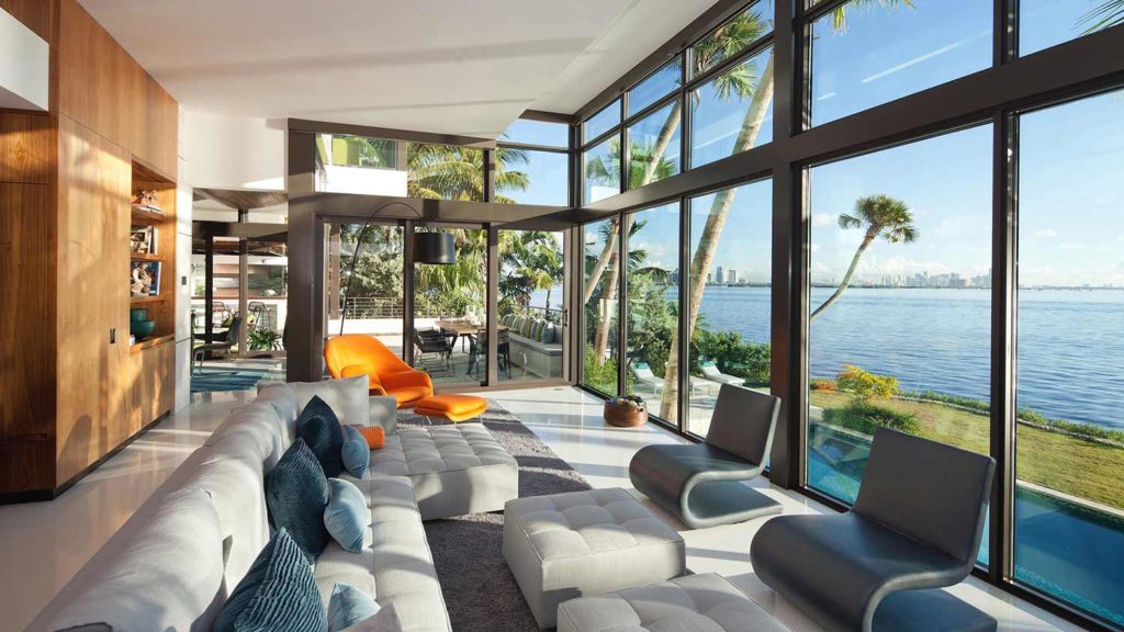 Touzet Studio drew inspiration from nature- the tropical hammock of Corlal Gables and Biscayne Bay beyond.

The House transitions from two engaged stone-clad volumes on the street to a primarily crystalline façade on the Bayside.

The interiors balance a warm and neutral palette with moments of intense color, utilizing terrazzo floors and natural wood to give the house a very Florida tropical feeling.

The blues and grey of the carpets and custom light fixtures pick up on the shades of the ocean. The bright bursts of color recall the vibrant colors found in the gardens of the Tropical landscapes.

The Furniture layout takes full advantage of the crystalline façade and beautiful bay views while also meeting the Clients programmatic needs.